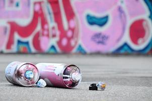 Several used spray cans with pink and white paint and caps for spraying paint under pressure is lies on the asphalt near the painted wall in colored graffiti drawings photo