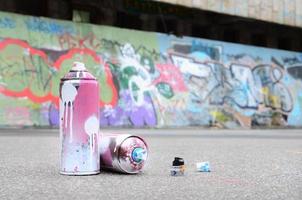 Several used spray cans with pink and white paint and caps for spraying paint under pressure is lies on the asphalt near the painted wall in colored graffiti drawings photo