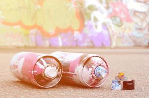 Several used spray cans with pink and white paint and caps for spraying paint under pressure is lies on the asphalt near the painted wall in colored graffiti drawings photo