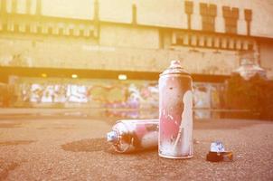 Several used spray cans with pink and white paint and caps for spraying paint under pressure is lies on the asphalt near the painted wall in colored graffiti drawings photo