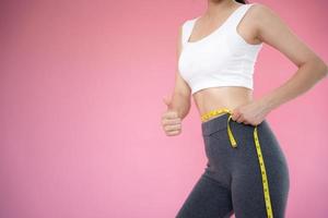 Slim woman in sportswear thumbs up and measures waist with tape measure on pink background. diet woman and achieve weight loss goals photo