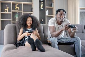 Father teases and pranks his daughter while playing console games on vacation. The black daughter and father smile happily together. Family activities on holidays. single father photo