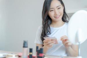 hermosa mujer asiática sentada frente a un espejo. cara de una mujer sana aplicando crema y maquillaje. anuncio de crema para la piel, crema antiarrugas, cara de bebé