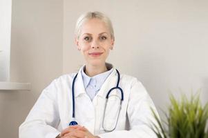 Portrait of smiling young Caucasian female doctor sit at desk in hospital or private clinic in white medical uniform at workplace, healthcare concept photo