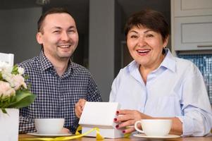 concepto de celebración navideña. sorprendida anciana sonriente sosteniendo un regalo sentada cerca de su hijo adulto. vacaciones de primavera. 8 de marzo, cumpleaños, día de la madre. foto