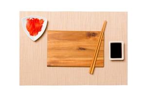 Empty rectangular brown wooden plate with chopsticks for sushi, ginger and soy sauce on brown sushi mat background. Top view with copy space for you design photo