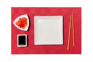 Empty white square plate with chopsticks for sushi and soy sauce, ginger on red mat sushi background. Top view with copy space for you design photo