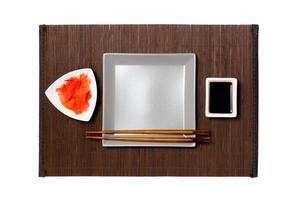 Empty gray square plate with chopsticks for sushi, ginger and soy sauce on dark bamboo mat background. Top view with copy space for you design photo