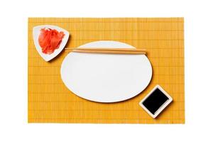 Empty oval white plate with chopsticks for sushi and soy sauce, ginger on yellow bamboo mat background. Top view with copy space for you design photo