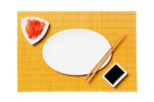 Empty oval white plate with chopsticks for sushi and soy sauce, ginger on yellow bamboo mat background. Top view with copy space for you design photo