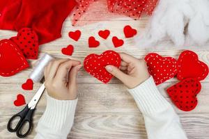 Valentine day theme. Workplace for preparing handmade decorations. Top view of female hands sew felt heart. Packed gifts, tools shabby wooden table photo