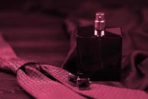 A bottle of mens cologne and cufflinks with blue tie lie on a black luxury fabric background on a wooden table. Mens accessories Image toned in Viva Magenta, color of the 2023 year photo