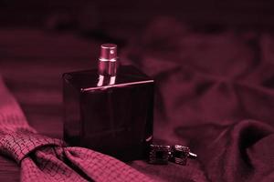A bottle of mens cologne and cufflinks with blue tie lie on a black luxury fabric background on a wooden table. Mens accessories Image toned in Viva Magenta, color of the 2023 year photo