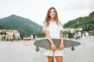 Slim young woman with long blonde hair in light sports clothes with longboard in outdoor skatepark at sunset photo