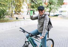 Portrait of fashionable happy young pretty woman in cap and sunglasses using mobile on bicycle on sunny autumn day in city park. Mobile mockup, smartphone with green screen photo