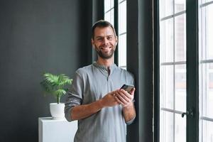 Young bearded man in gray longsleeve with smartphone in hands reading message standing at window in modern office with dark walls photo