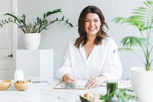 joven morena sonriente doctora nutricionista de talla grande con camisa blanca trabajando en una laptop en una oficina moderna y luminosa. el médico prescribe una receta de medicamentos y vitaminas en la clínica foto