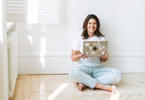 mujer morena sonriente con ropa informal de talla grande con cuerpo positivo usando una laptop sentada en el suelo en la habitación luminosa foto