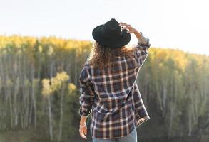 Young beautiful woman with curly hair in felt hat and plaid shirt in jeans looks at magic view of mountains and river, hiking on autumn nature photo