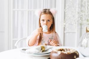 una niñita linda y graciosa con el pelo largo y un vestido rosa claro bebiendo té en una mesa festiva en un salón luminoso en casa. tiempo de navidad, cumpleañera foto