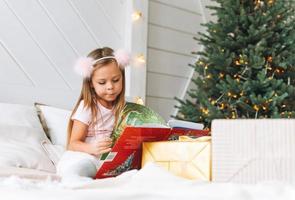 linda niñita con vestido rosa leyendo un libro sentada en la cama en la habitación con un árbol de navidad. niño en llanta con cuernos de ciervo en casa de campo en Navidad, feliz año nuevo foto