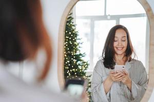 Young adult forty years middle age beautiful woman with dark hair in cozy pajamas looking at mirror and taking selfie on the Christmas morning in room at the home photo