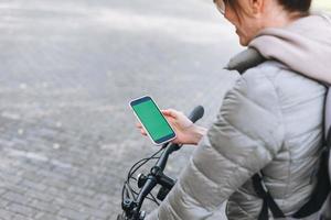 Portrait of fashionable happy young pretty woman in cap and sunglasses using mobile on bicycle on sunny autumn day in city park. Mobile mockup, smartphone with green screen photo