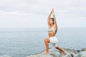 joven mujer hermosa en forma con cabello largo en ropa deportiva blanca practica yoga y disfruta de la vida en la playa del mar, salud mental y física foto