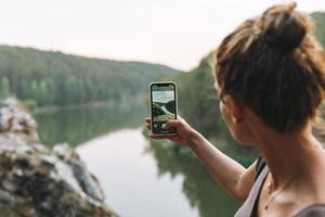 joven mujer delgada con mochila tomando fotos de la hermosa vista de las montañas y el río tranquilo en el teléfono móvil, gente de atrás, viajes locales