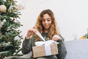 Teenage girl with curly hair holds gift box in her arms while sitting on the sofa in the living room with a Christmas tree. Happy teenager in the decorated living room at house on Christmas morning photo