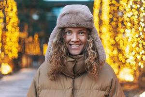 Portrait of young smiling woman with curly hair in fur hat in the winter street decorated with lights photo