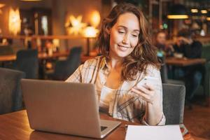 Encantadora mujer morena adulta con camisa a cuadros que trabaja con una laptop usando un teléfono móvil en el café foto