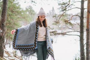 joven morena hermosa mujer con sombrero y poncho gris en el bosque de invierno foto