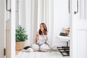 Young adult forty years beautiful woman with long hair in cozy pajamas using mobile smartphone sitting on floor in room with Christmas tree at the home photo