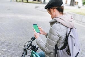 Portrait of fashionable happy young pretty woman in cap and sunglasses using mobile on bicycle on sunny autumn day in city park. Mobile mockup, smartphone with green screen photo