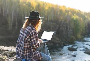 Young beautiful woman with curly hair in felt hat and plaid shirt using laptop on magic view background of mountains and river, hiking on autumn nature, concept of freelance photo