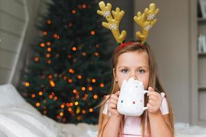 Cute smiling funny little girl in pink dress drinking cocoa sitting on bed in room with Christmas tree. Portrait of child in the rim with deer horns in cottage house in christmas time, happy New Year photo