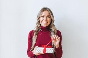 Portrait of happy young adult beautiful woman with blonde long curly hair in bordo sweater with gift box red ribbon in hands on grey background isolated, winter warm clothes photo