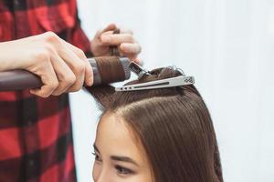 Hair stylist barber styling long hair with hair iron for beautiful young asian woman with cup of tea in beauty salon, working moment photo