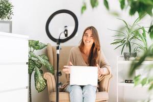 Young pretty woman blogger records video on mobile phone using ring lamp sitting on chair with laptop in room with green plants at home photo