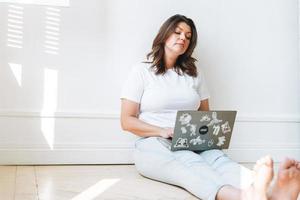 mujer morena sonriente con ropa informal de talla grande con cuerpo positivo usando una laptop sentada en el suelo en la habitación luminosa foto