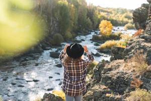 Young beautiful woman with curly hair in felt hat and plaid shirt in jeans looks at magic view of mountains and river, hiking on autumn nature photo