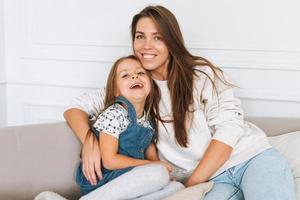 retrato de una joven familia feliz en un sofá en un luminoso salón. mujer feliz con una linda niña sonriendo y mirando a la cámara. ocio conjunto en casa foto