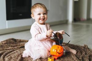 Cute little baby girl in pink dress with the witch's hat and pumpkin lantern sitting on floor in kitchen at the home, Halloween time photo