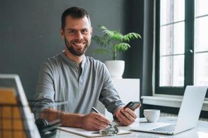 Young smiling business man in casual grey longsleeve working at laptop in dark modern office room photo