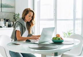 joven adulta de cuarenta años hermosa mujer rubia con el pelo largo trabajando en una laptop sentada en la cocina en casa, trabajo remoto foto