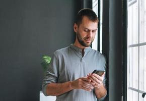 Young bearded man in gray longsleeve with smartphone in hands reading message standing at window in modern office with dark walls photo