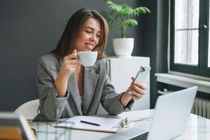 joven morena sonriente con el pelo largo y traje gris usando un teléfono móvil trabajando en una laptop en la oficina moderna foto