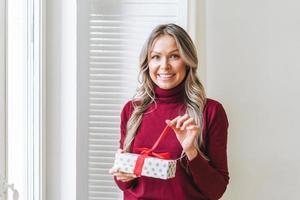 Portrait of happy young adult beautiful woman with blonde long curly hair in bordo sweater with gift box red ribbon in hands near window in the bright interior, winter warm clothes photo