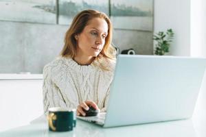 joven adulta de cuarenta años hermosa mujer rubia con el pelo largo trabajando en una laptop sentada en la cocina en casa, trabajo remoto foto